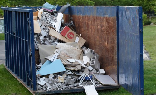 A large industrial skip container, situated outdoors on a grassy area typical of Rotherhithe, filled with a mixture of construction and household waste. The contents include broken pieces of drywall, plasterboard, tiles, and concrete fragments, alongside cardboard boxes, plastic, and metal debris. The skip's exterior is dark blue, with visible rust and wear on the inner walls, indicating frequent use for rubbish removal. The surrounding background includes a neatly maintained lawn and trees, with overhanging greenery suggesting a park or garden setting. The waste appears to be recently collected and is piled up to the top of the container, with some materials spilling over the edges. The scene reflects a typical rubbish disposal process managed by firms like Commercial Waste Rotherhithe, who handle waste clearing and recycling from local premises. The image emphasizes the various textures and colors of the materials, from rough concrete to smooth cardboard and shiny plastic, capturing the nature of commercial waste management in the area.