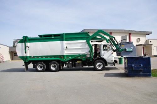 A professional rubbish collection vehicle with a white cargo container and green structural accents, positioned on a paved street in a suburban area. The truck features a hydraulic lift arm at the front of the cargo area, designed for lifting and emptying waste bins. It is parked near a dark blue wheelie bin placed on the street, with an additional dark green bin situated on the curb. The surroundings include modern residential buildings with flat roofs and clean architectural lines, typical of Rotherhithe. The scene is captured during daytime with overcast skies, providing natural, diffuse lighting that highlights the vehicle's clean exterior and the textures of the bin surfaces. This image visually supports rubbish removal services offered by Commercial Waste Rotherhithe, illustrating their waste collection process within a local urban environment.