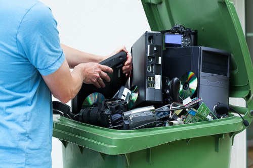 A person wearing a light blue t-shirt is holding a black electronic device over an open green waste bin filled with discarded electronic items, including computer towers, circuit boards, keyboards, and CDs, all of which are visibly stacked and spilling over the top. The electronic waste features a mix of black, silver, and green components, with textures ranging from glossy plastic to metallic surfaces. The environment appears to be outdoors, possibly on a driveway or paved area, with natural light illuminating the scene. This image illustrates a waste collection process for electronic or rubbish removal, relevant to companies like Commercial Waste Rotherhithe operating within the Rotherhithe area, perhaps in South East London, where electronic waste disposal is part of comprehensive rubbish management services.