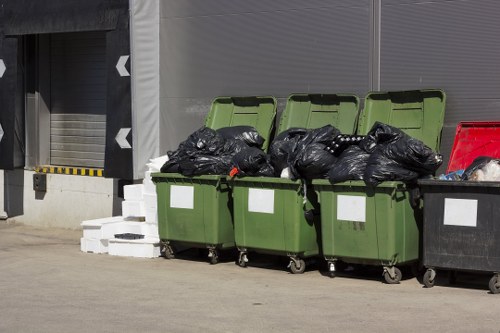 A row of four large wheelie bins positioned on a concrete surface outside a commercial building, with the first three bins in green and the fourth in black. The green bins have their lids partially open, revealing black rubbish bags stacked inside, some protruding from the lids. The black bin appears to be filled with waste, with its lid slightly ajar. Behind the bins, there is a grey industrial wall with a partially visible roll-up door and a small section of a loading bay. To the left, a stack of white bricks or blocks is placed on the ground. The scene is evenly lit, indicating natural daylight, and the environment suggests a typical waste disposal area in a commercial setting, suitable for rubbish removal services in Rotherhithe or nearby areas.