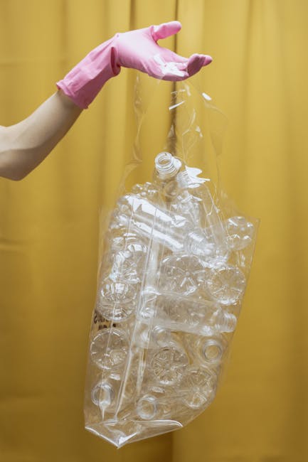 A person wearing a light pink rubber glove is holding a large, clear plastic bag filled with numerous transparent plastic bottles, some crushed and others intact, against a plain yellow background. Th